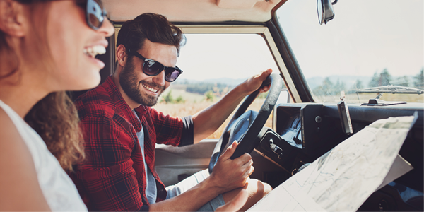 A man driving a car smiles as he looks over at a laughing woman in the passenger seat holding a paper roadmap.